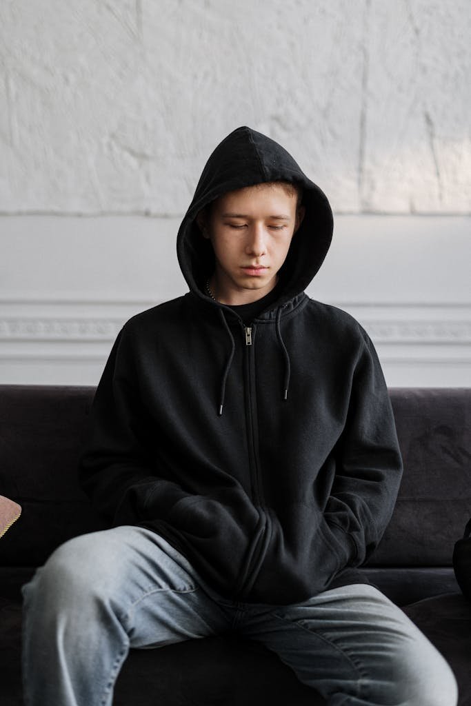 Teenager in a black hoodie sitting on a couch, reflecting on thoughts indoors.